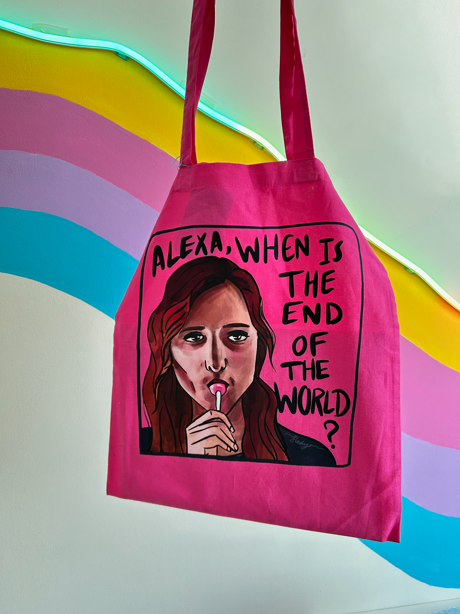 Pink tote bag with a graphic of a woman and text 'ALEXA, WHEN IS THE END OF THE WORLD?' against a colorful background.